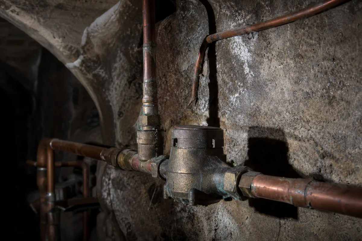 Cave voûtée d&rsquo;une maison ancienne en pierres dorées, canalisation de cuivre et compteur d&rsquo;eau sous une lumière technique contrastée