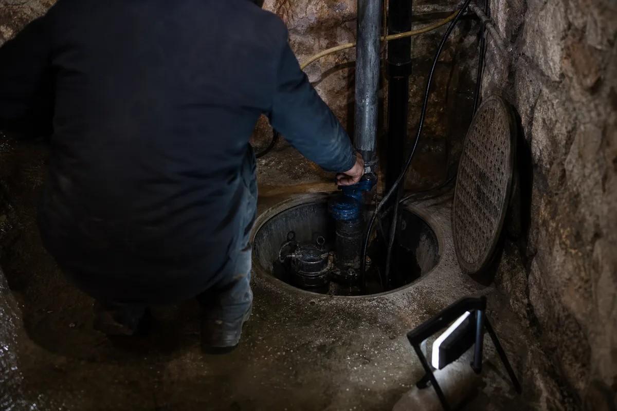 Plombier de dos manœuvrant la vanne d&rsquo;une pompe de relevage dans un poste de cave, lumière technique contrastée