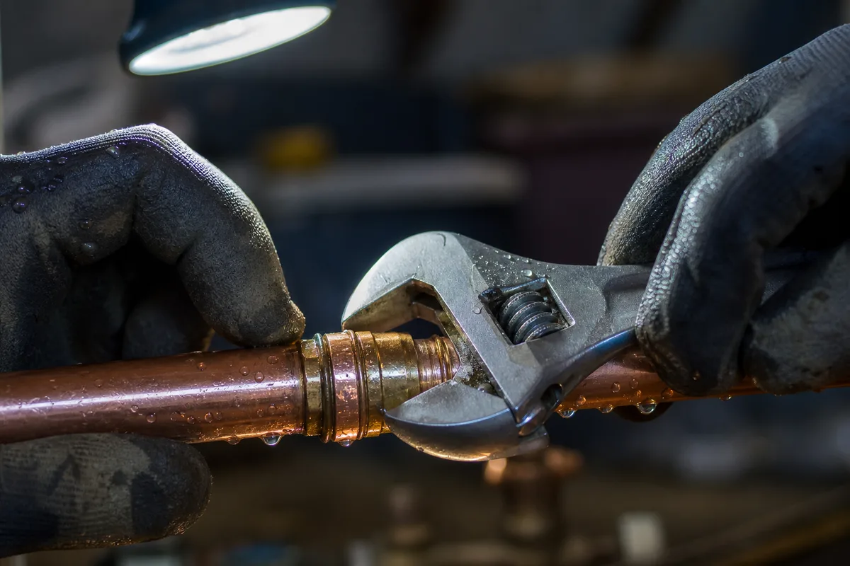 Gros plan sur des mains serrant un raccord en cuivre à la clé sous une lampe de travail, gouttes d’eau